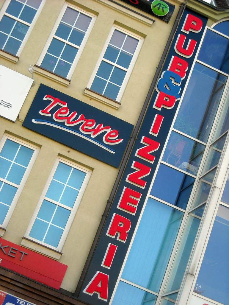 Zewnętrzny widok budynku z szyldami 'Tevere' i 'Pub & Pizzeria' w czerwono-niebieskiej kolorystyce, widoczne okna i przeszklona klatka schodowa.