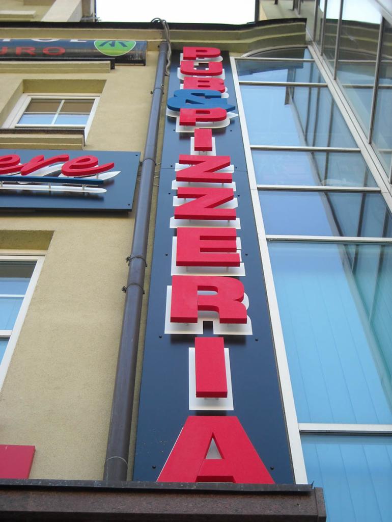 Pionowy szyld z nazwą 'Pizzeria' w kolorze czerwono-niebieskim zamontowany na fasadzie budynku, widok z dołu.