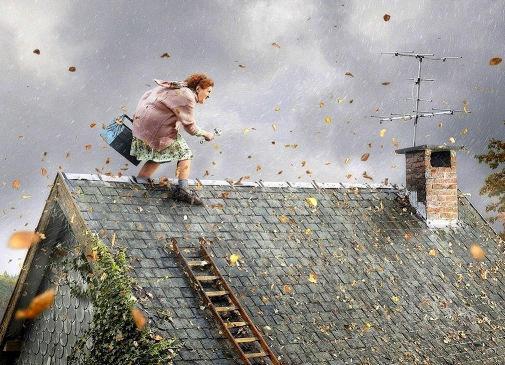 Kobieta z torbą i narzędziem na dachu domu podczas wietrznej pogody, w tle komin i antena telewizyjna.