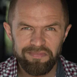 Portret mężczyzny z brodą, w koszuli w kratę i naszyjniku z koralików, o przenikliwym spojrzeniu.