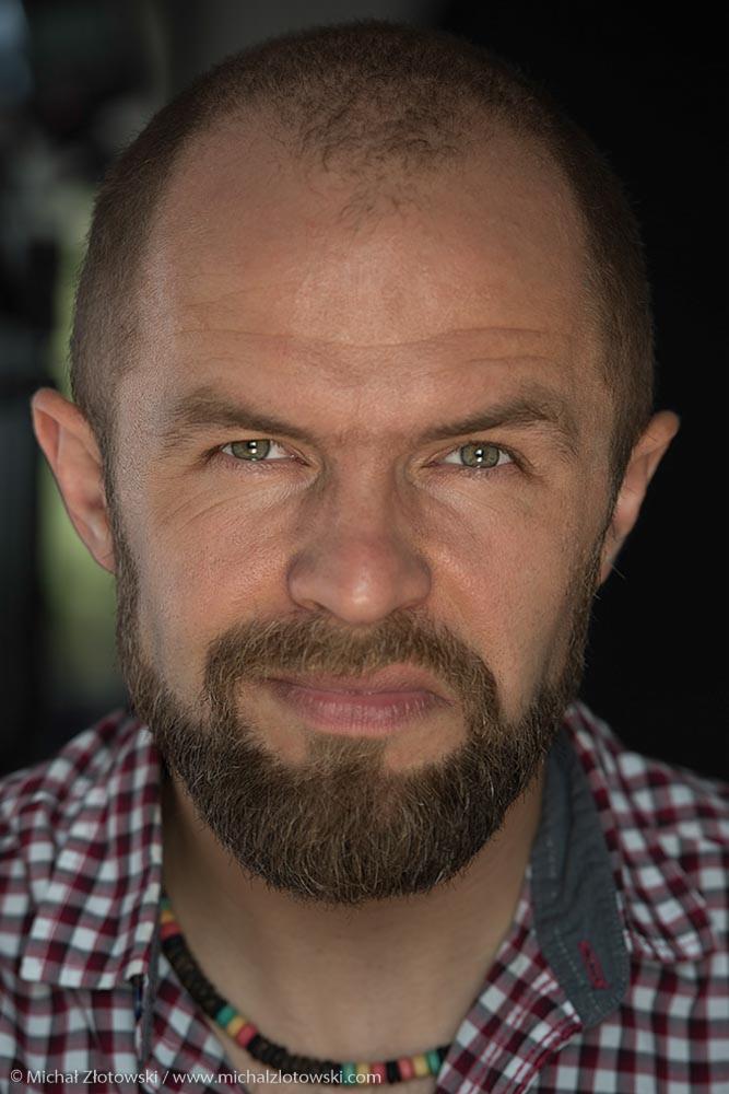 Portret mężczyzny z brodą, w koszuli w kratę i naszyjniku z koralików, o przenikliwym spojrzeniu.