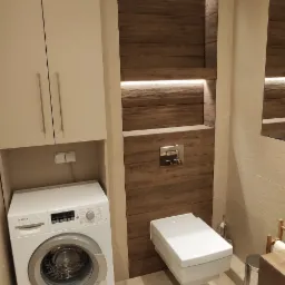 Nowoczesna łazienka z podwieszaną toaletą, pralką Bosch i oświetloną wnęką z drewnopodobnymi panelami.