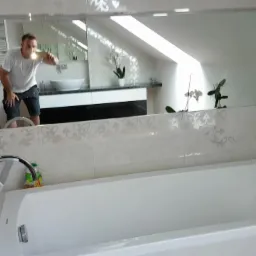 Wykończona, jasna łazienka z białą wanną, chromowaną armaturą i lustrem w którym odbija się osoba robiąca zdjęcie, widoczna umywalka i okno dachowe.