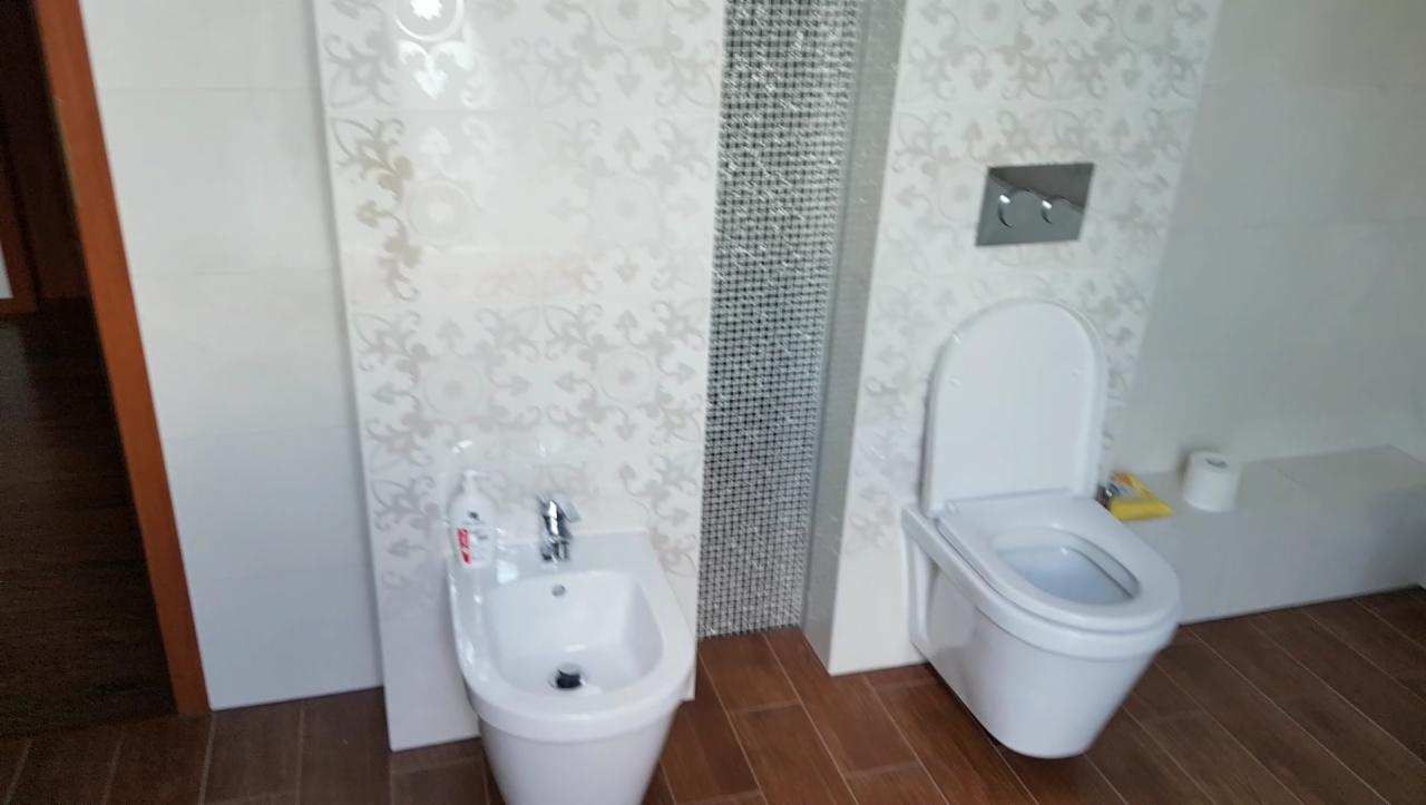 Biała łazienka z toaletą podwieszaną i bidetem, dekoracyjne płytki ścienne z motywem roślinnym i mozaiką, brązowa podłoga imitująca drewno.