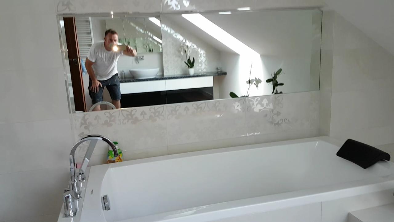 Wykończona, jasna łazienka z białą wanną, chromowaną armaturą i lustrem w którym odbija się osoba robiąca zdjęcie, widoczna umywalka i okno dachowe.