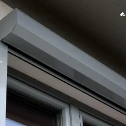 Szara roleta zewnętrzna opuszczona do połowy, zamontowana na oknie, widoczne logo 'bizmet' w prawym górnym rogu, ściana budynku w tle.