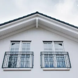 Dwa białe okna z czarnymi balustradami na białej fasadzie domu pod stromym dachem z ciemną dachówką, widok z dołu.