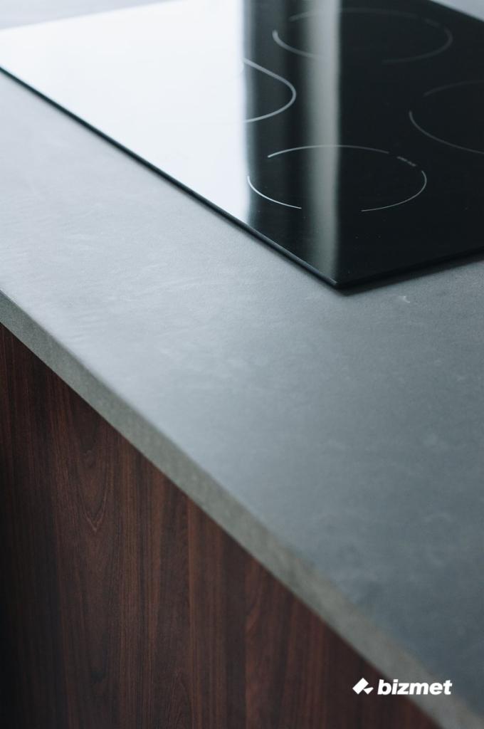 Minimalistyczny blat kuchenny z szarego kamienia z wbudowaną czarną płytą indukcyjną, wykończony ciemnym drewnem.