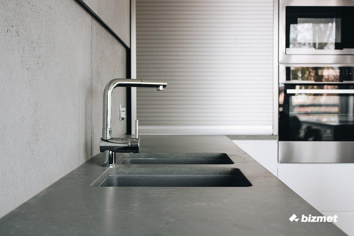 Nowoczesna kuchnia z szarym blatem, dwukomorowym zlewem i chromowaną baterią, w tle piekarnik i roleta okienna.