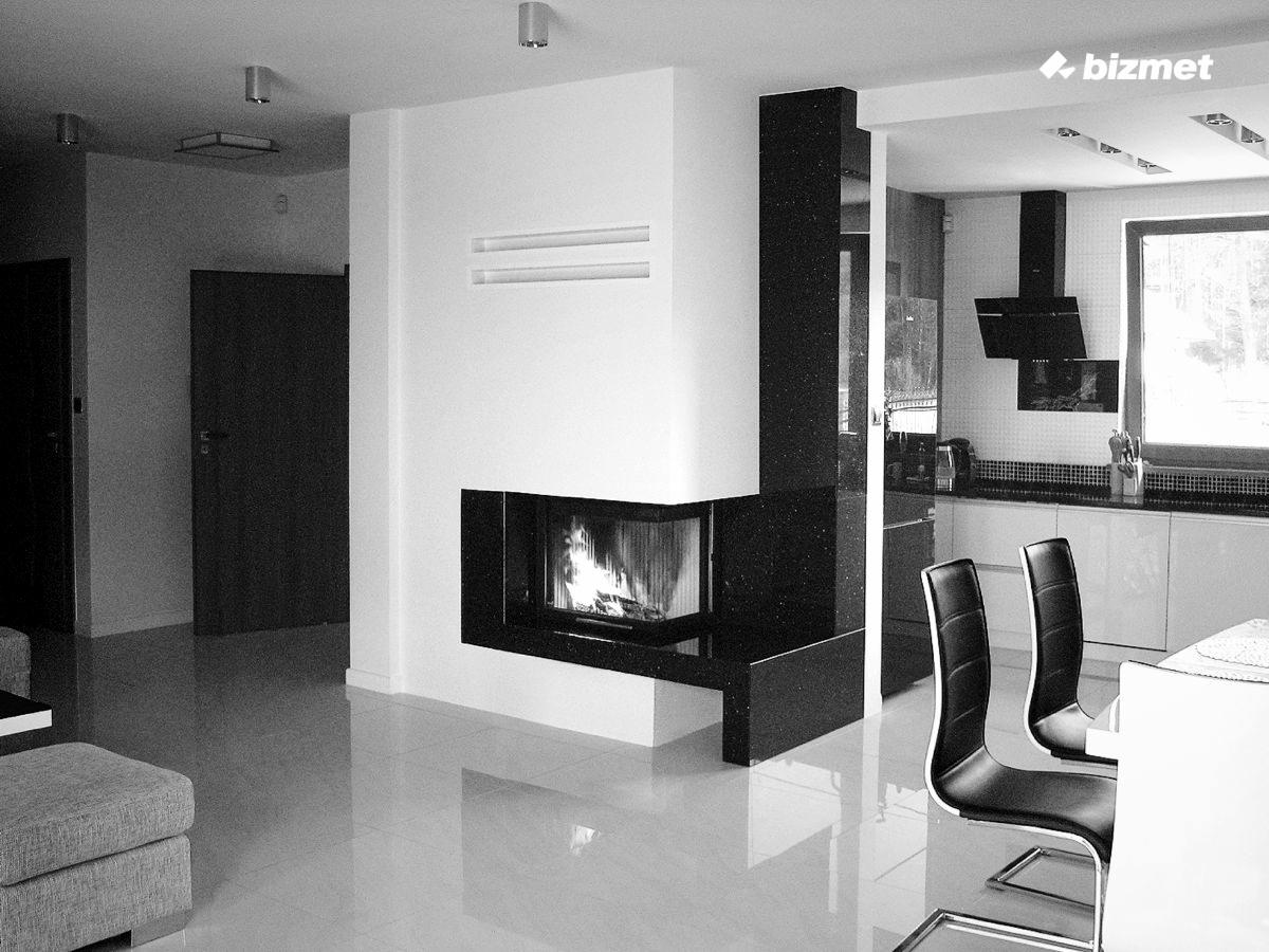 Czarno-białe wnętrze z nowoczesnym kominkiem narożnym, widok na kuchnię z okapem, minimalistyczne krzesła przy stole, połyskująca podłoga.