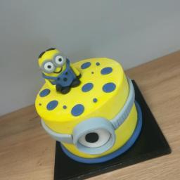 Pastelove - Żółty tort w stylu Minionka z niebieskimi akcentami, umieszczony na czarnej podstawie na drewnianym blacie.