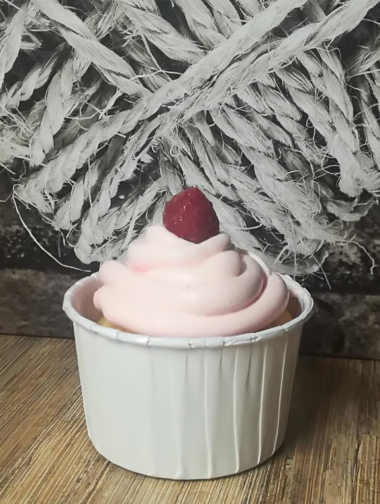 Babeczka w białej papilotce z różowym kremem i maliną na wierzchu, na drewnianym blacie, z tłem imitującym zwój liny.