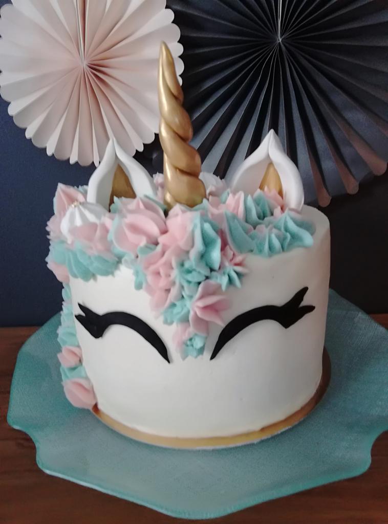 Tort jednorożec z białym kremem, złotym rogiem, pastelowymi różowymi i turkusowymi dekoracjami z kremu, na szklanym talerzu, z dekoracjami papierowymi w tle.
