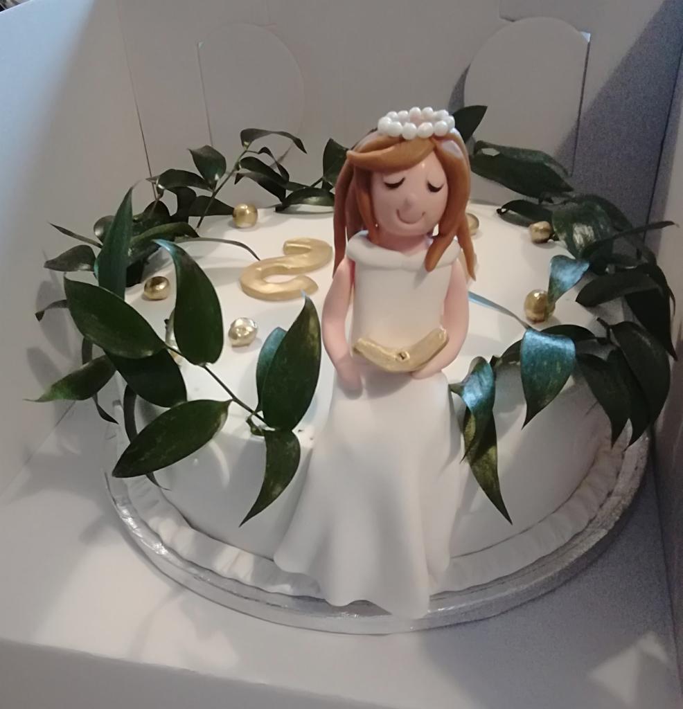 Tort komunijny z figurką dziewczynki w białej sukience, dekorowany zielonymi gałązkami i złotymi kuleczkami, umieszczony w białym pudełku.