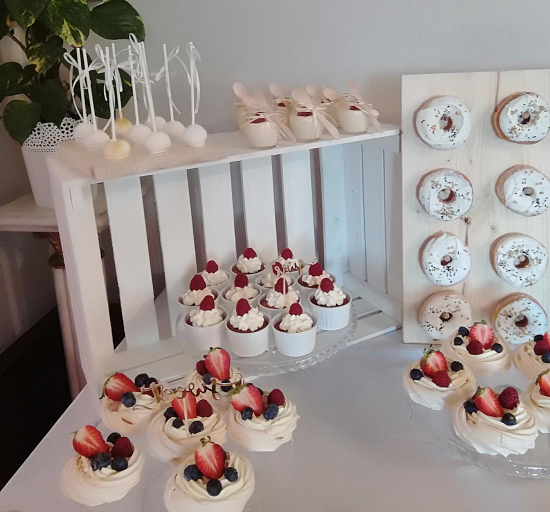 Stół z różnorodnymi deserami: mini bezy z owocami, babeczki z kremem i malinami, donuty na drewnianej desce, cake pops i desery w słoiczkach, ustawione na białym tle z rośliną doniczkową w tle.