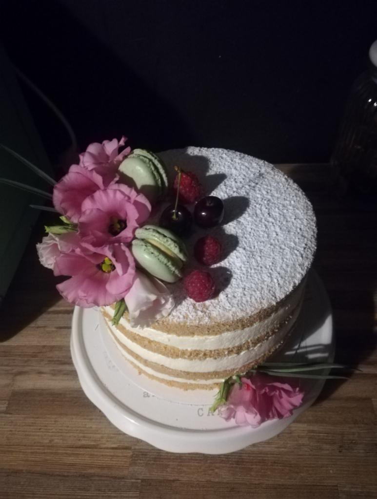 Tort warstwowy z kremem, posypany cukrem pudrem, udekorowany różowymi kwiatami, malinami, wiśniami i zielonymi makaronikami, ustawiony na białym talerzu na drewnianym stole.