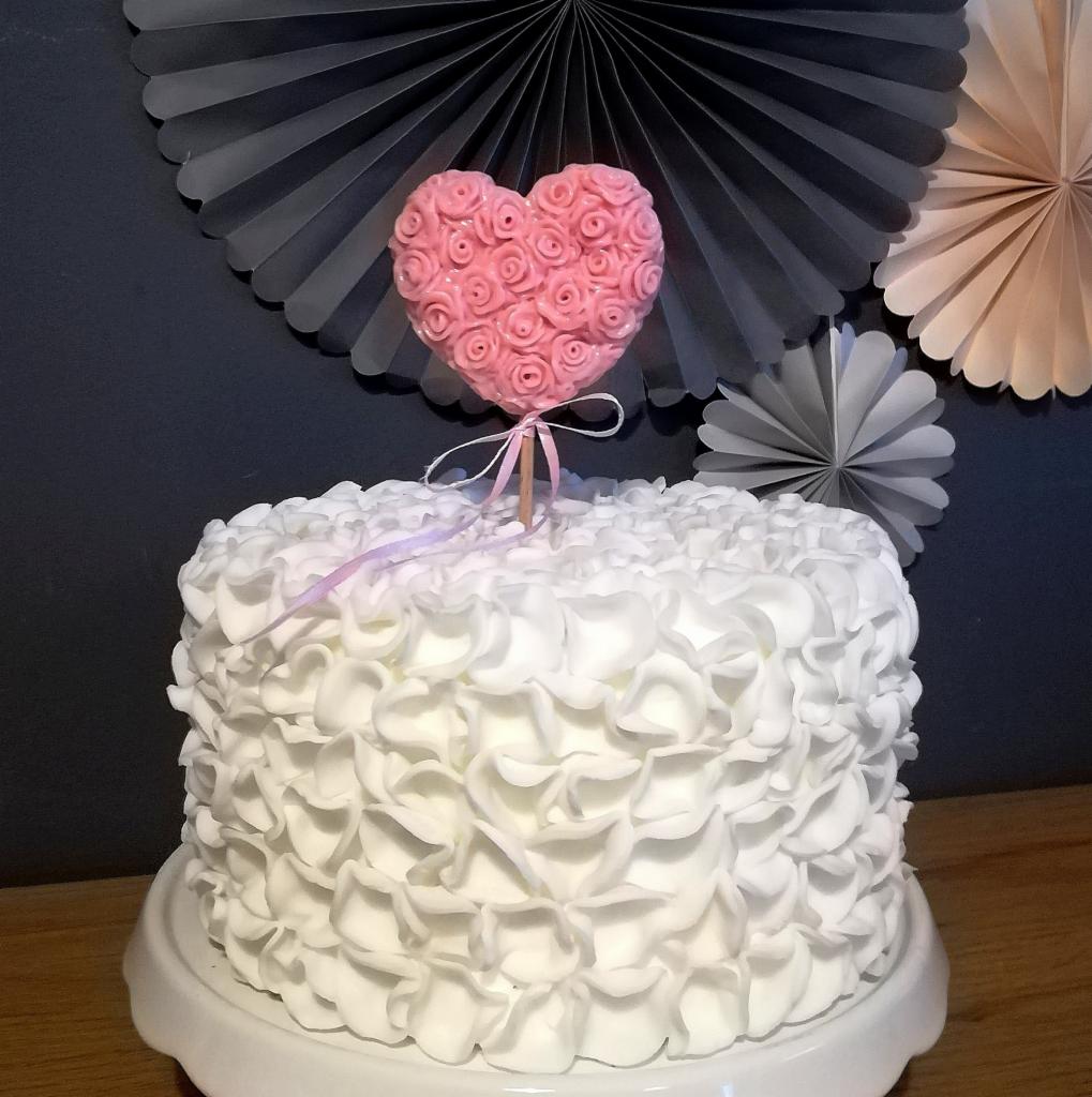 Tort z białym kremem w stylu ruffle cake, ozdobiony różowym sercem z różyczek na patyku, przewiązanym wstążką, na tle dekoracyjnych rozet; wykonany w cukierni w Lesznie.
