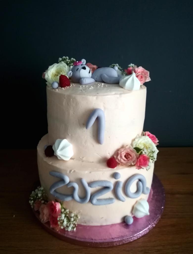 Dwupoziomowy tort w odcieniach różu, udekorowany kwiatami, bezami, malinami, figurką śpiącego misia z lukru i napisem 'Zuzia' z lukru, z cyfrą 1 na górnym poziomie.