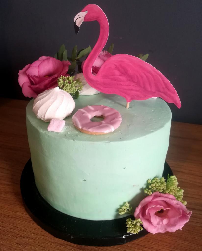 Tort z jasną, miętową polewą, udekorowany różowym flamingiem na patyku, różami, bezą i pączkiem z różowym lukrem, na drewnianym blacie.