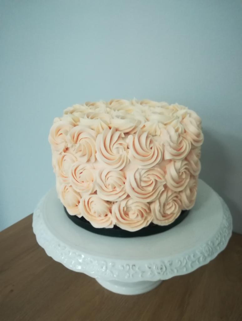 Tort z kremem w kształcie róż na białym paterze, tło jasnoniebieskie.