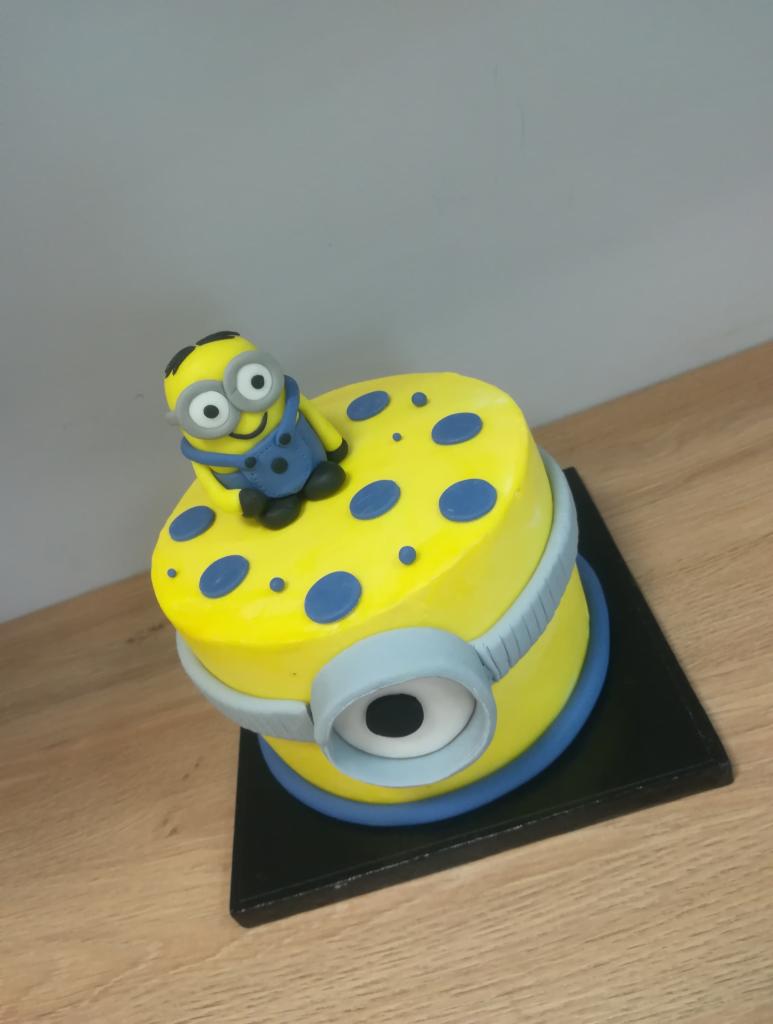 Żółty tort w stylu Minionka z niebieskimi akcentami, umieszczony na czarnej podstawie na drewnianym blacie.