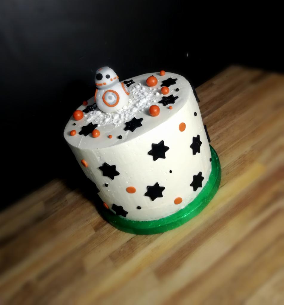 Tort z białym kremem, czarnymi gwiazdami i pomarańczowymi kulkami, zwieńczony figurką robota BB-8 na posypce imitującej śnieg, umieszczony na zielonym podkładzie.