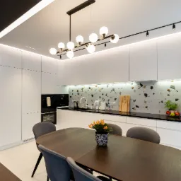 Nowoczesna kuchnia z białymi szafkami, czarnym blatem i mozaikową ścianą, oświetlona lampami kulistymi, z ciemnym, rozkładanym stołem i szarymi krzesłami.