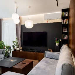 Nowoczesny salon z szarą sofą, czarnym panelem TV, drewnianą ścianą lamelową i wiszącymi lampami kulami.