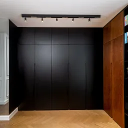 Czarna zabudowa meblowa od podłogi do sufitu z ukrytymi drzwiami, oświetlenie szynowe sufitowe, parkiet w jodełkę, obok regał z drewnianymi frontami i czarnymi półkami z książkami.