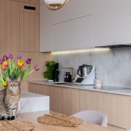 FURSTA Meble Wojciech Ziembla - Jasna kuchnia z drewnianymi frontami, kwiatami i kotem siedzącym na stole. Nowoczesny design, oświetlenie LED pod szafkami, sprzęt AGD na blacie.