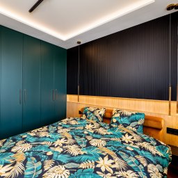 FURSTA Meble Wojciech Ziembla - Nowoczesna sypialnia z szafą w zabudowie w odcieniach zieleni, drewnianym wezgłowiem i czarną ścianą z pionowymi panelami. Nad łóżkiem wiszące, złote lampy.