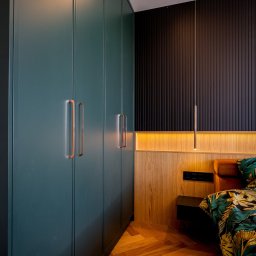 FURSTA Meble Wojciech Ziembla - Gładka, ciemnozielona szafa wnękowa z miedzianymi uchwytami w sypialni z drewnianą podłogą w jodełkę i czarną ścianą z lameli.