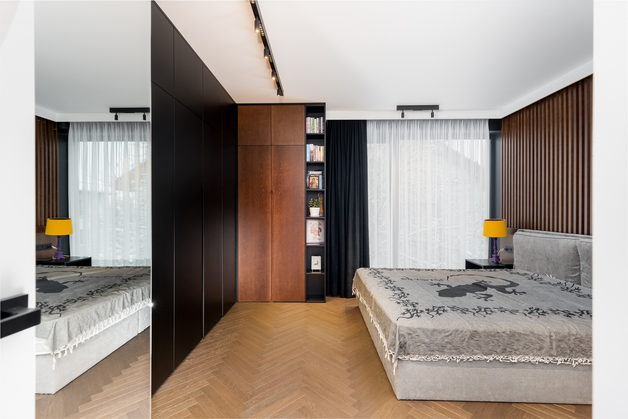 Nowoczesna sypialnia z dużą szafą w zabudowie, łączącą czarne fronty z drewnianymi, oraz regałem na książki, uzupełniona o drewnianą boazerię i parkiet w jodełkę.