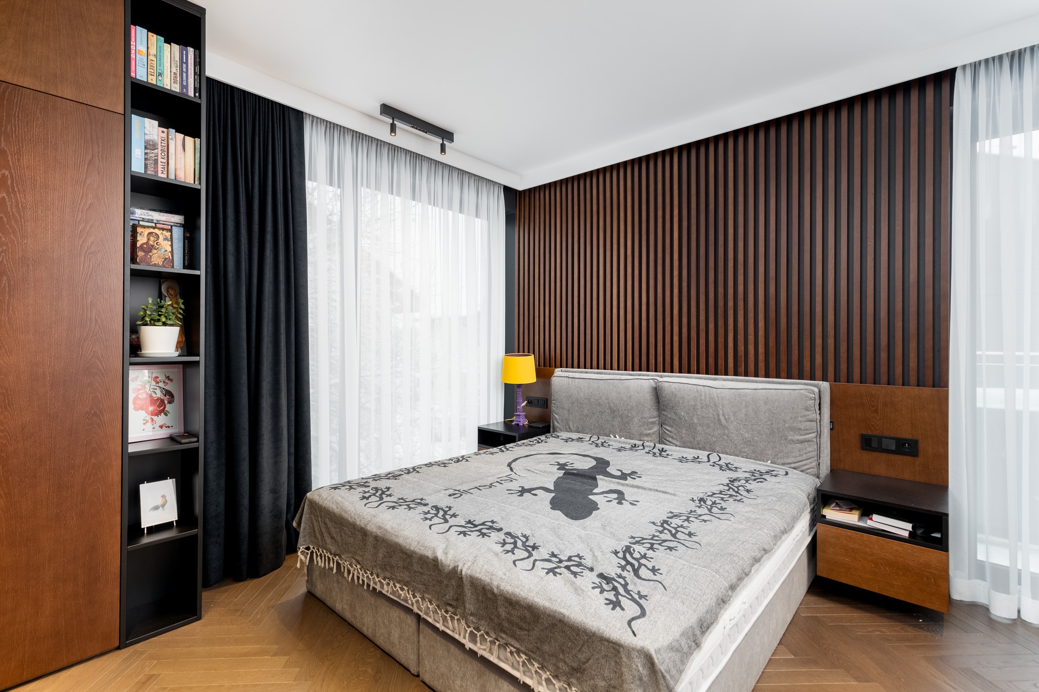 Nowoczesna sypialnia z drewnianą ścianą, łóżkiem z szarą narzutą w jaszczurki, drewnianą podłogą w jodełkę i regałem na książki w zabudowie.