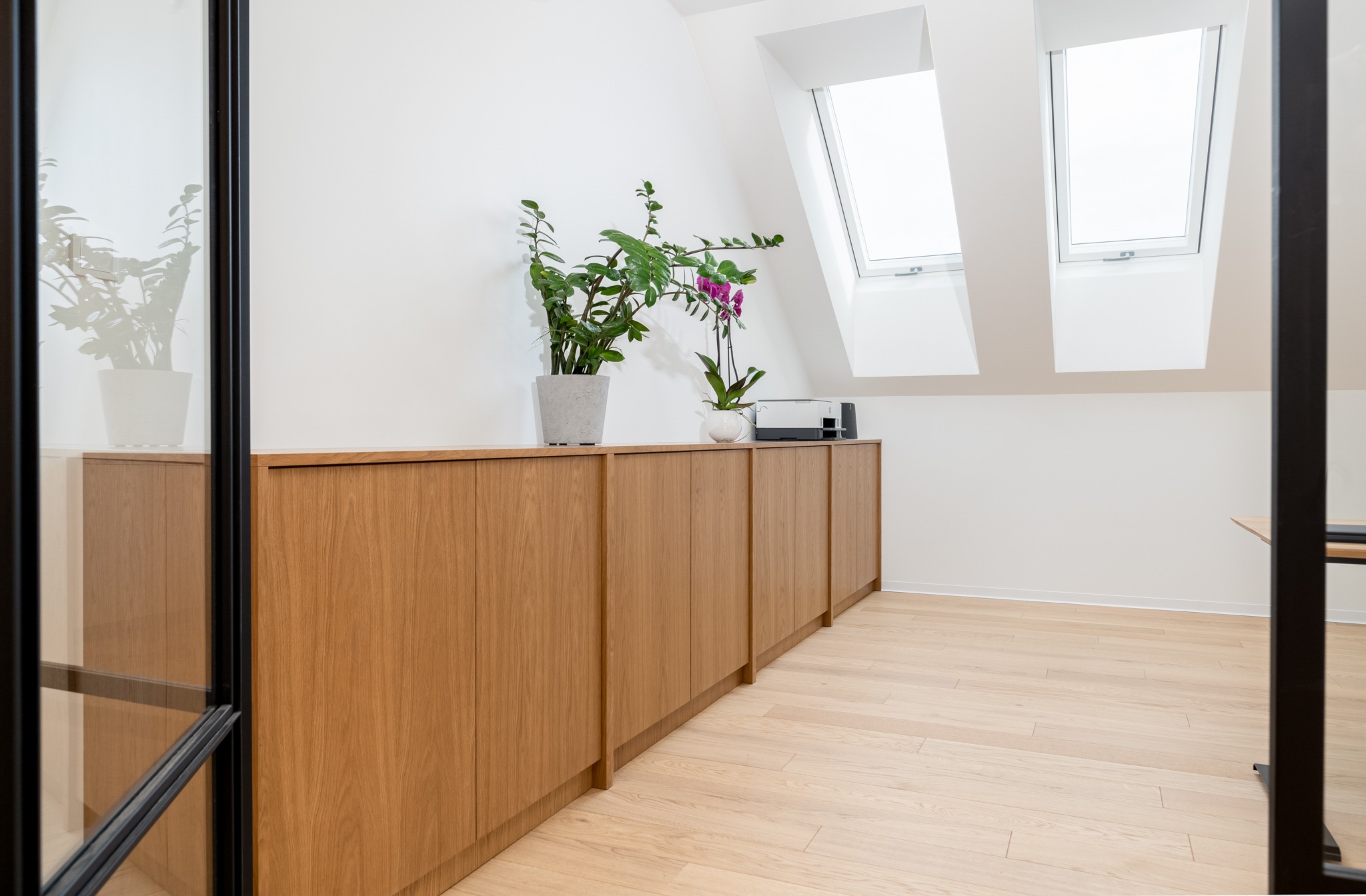 Długa, niska zabudowa meblowa z drewnianymi frontami pod skosami dachu, na blacie doniczki z zielonymi roślinami i storczykiem, widok zza szklanej ściany z czarnymi szprosami.