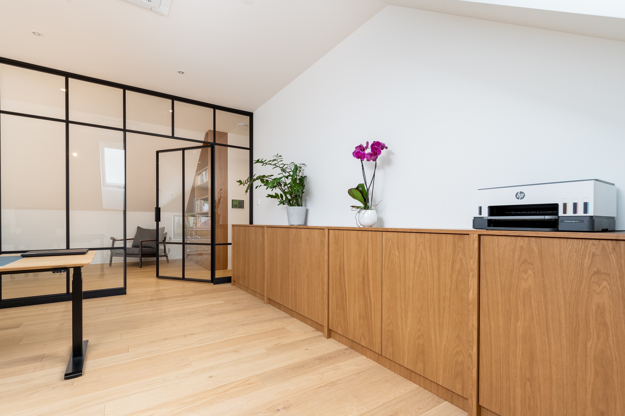 Jasne biuro z drewnianą podłogą i szafkami, przeszklona ściana z czarnymi ramami, biurko z regulowaną wysokością, drukarka HP i rośliny doniczkowe na szafkach.
