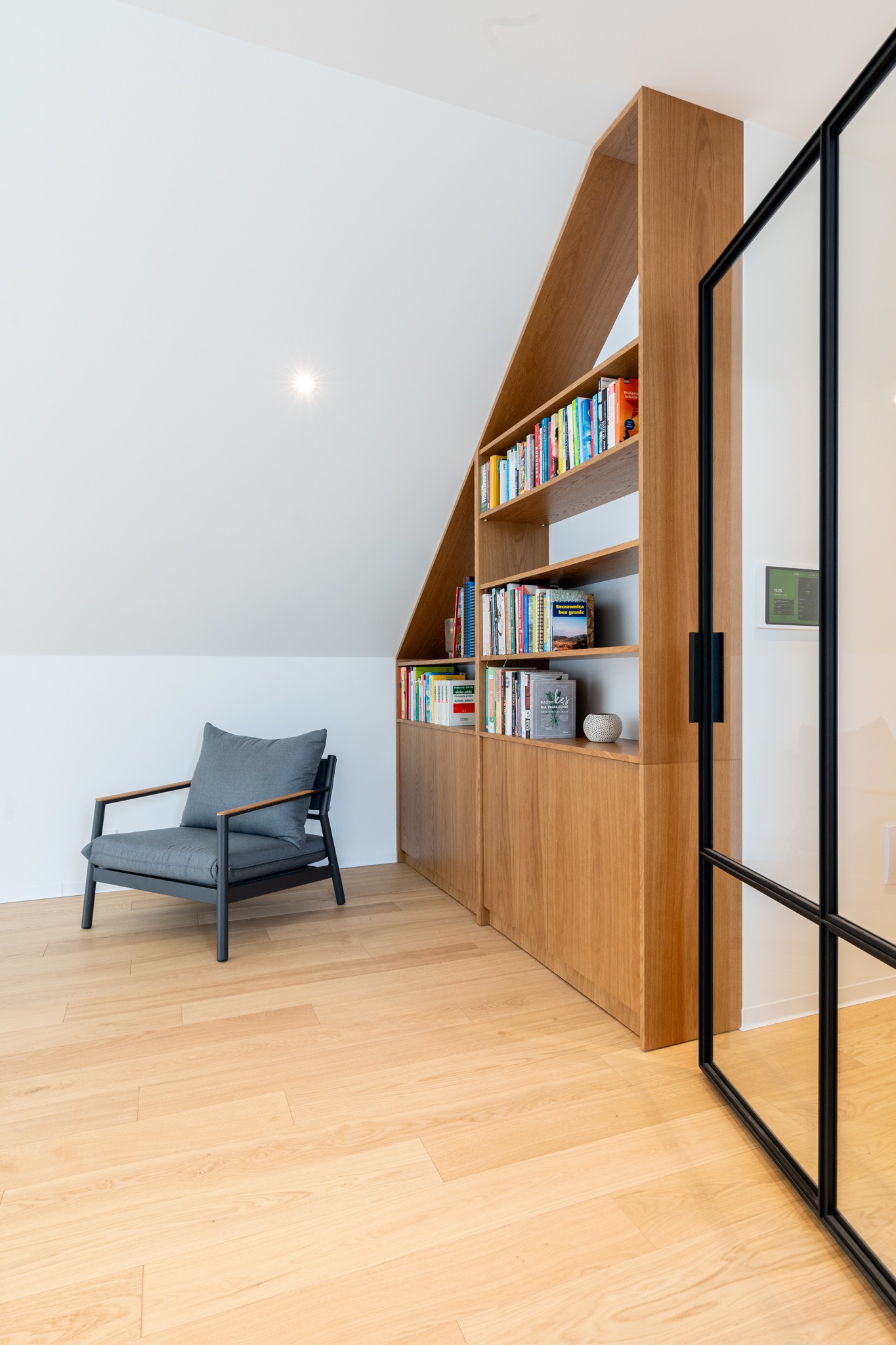 Drewniana biblioteczka na wymiar zabudowana pod skosem dachu, z książkami i dekoracjami, obok szary fotel i przeszklone drzwi z czarną ramą.