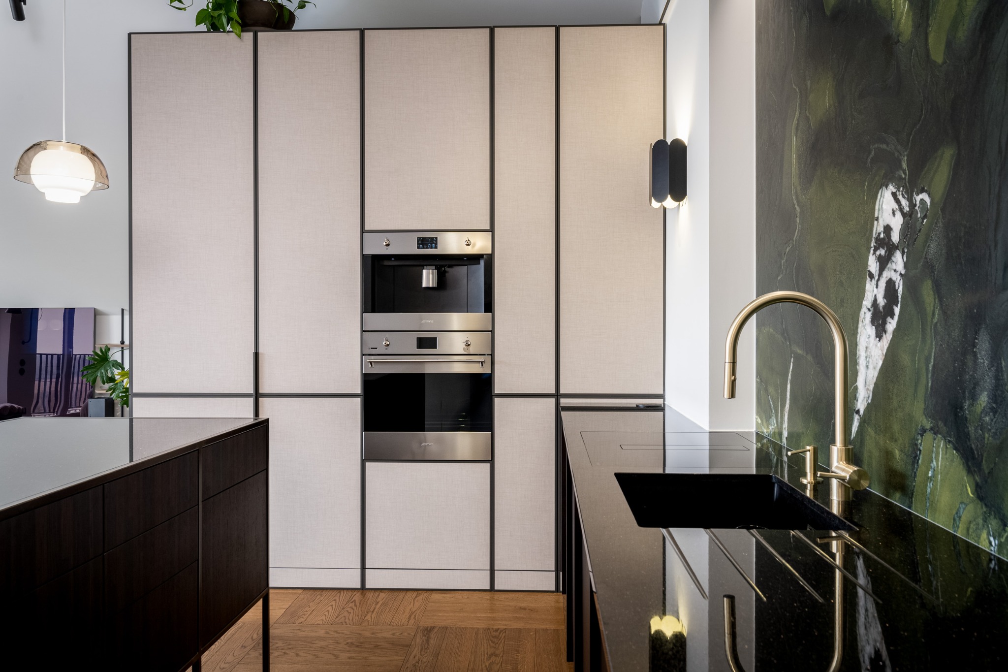 Elegancka kuchnia z zabudową w odcieniach beżu i czerni. W tle marmurowa ściana i złota bateria. Widoczny ekspres do kawy i piekarnik w zabudowie.