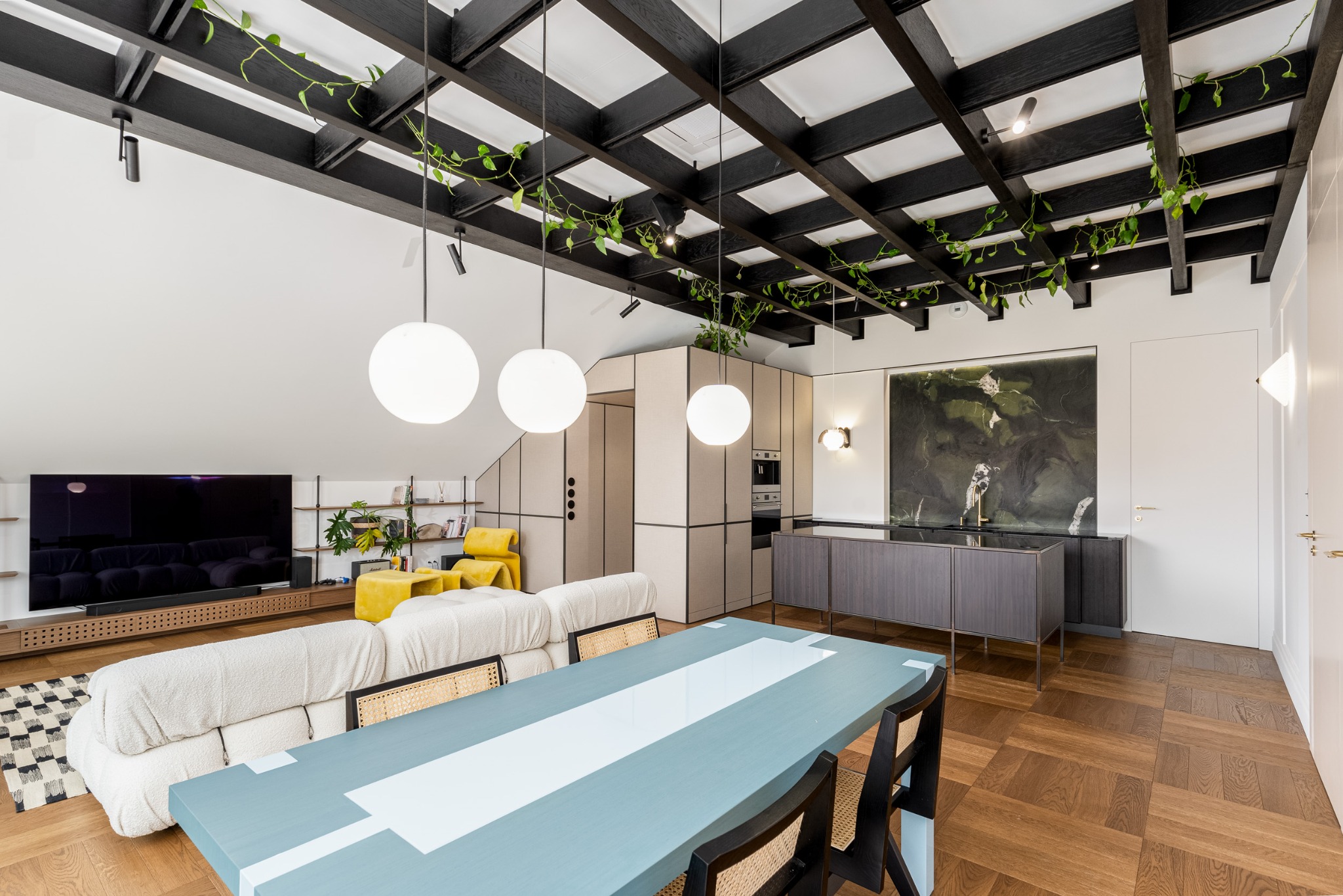 Nowoczesne wnętrze: stół z błękitnym blatem, kremowa sofa, zabudowa kuchenna z jasnymi frontami i czarnymi akcentami, drewniany parkiet, rośliny pnące się po suficie.
