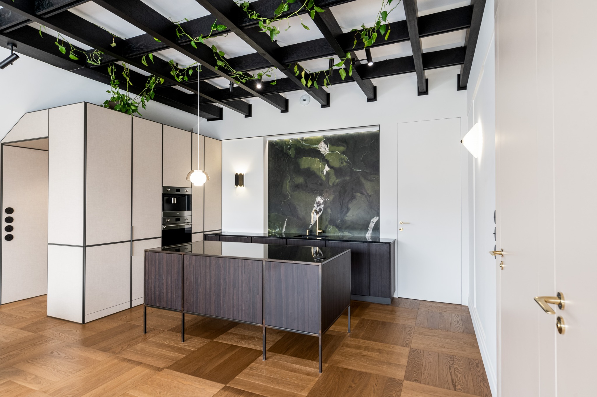 Nowoczesna kuchnia z wyspą na tle ściany z zielonego marmuru. Czarna, drewniana konstrukcja sufitowa z roślinami. Parkiet w jodełkę. Minimalistyczny design.