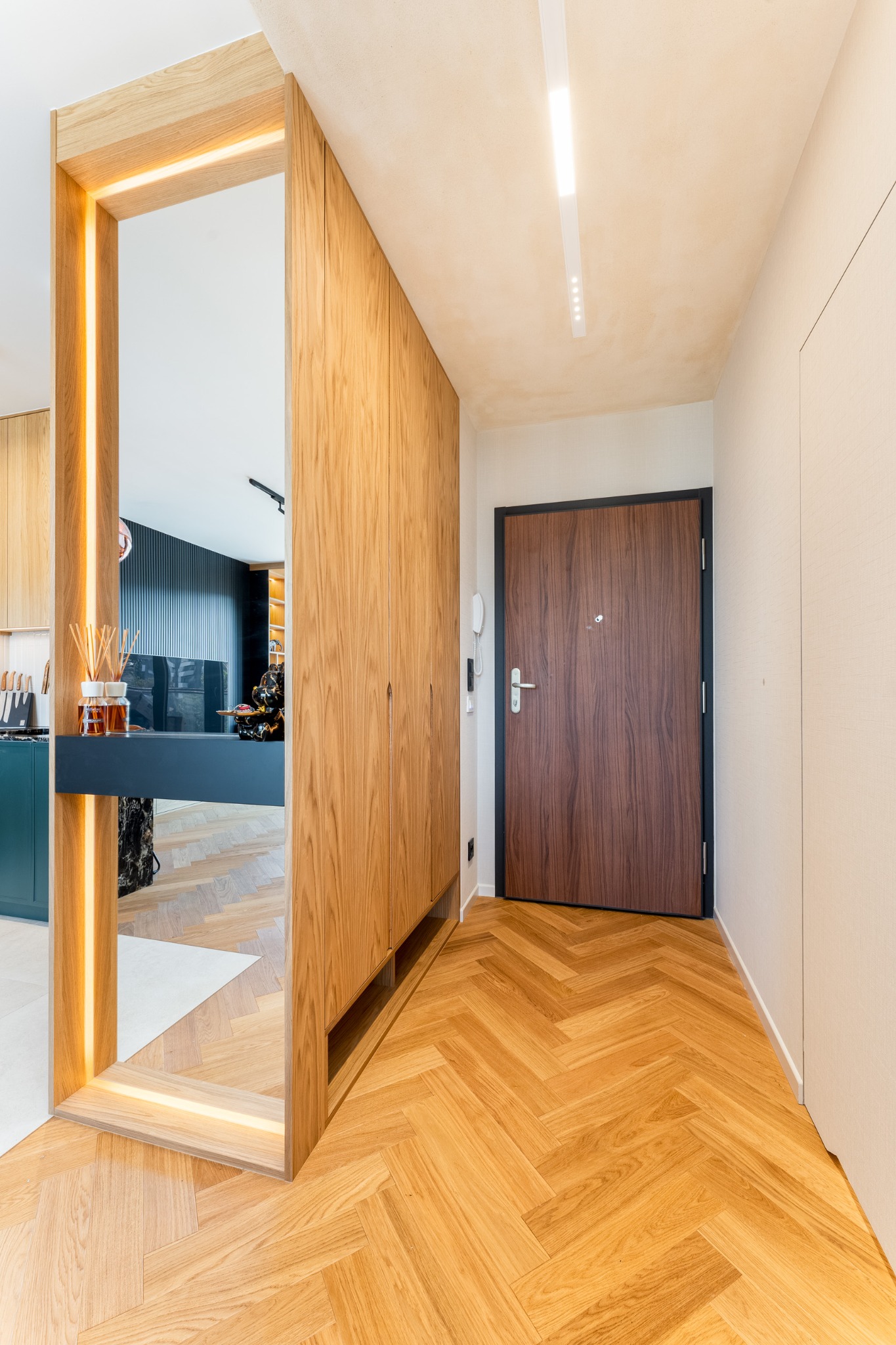 Nowoczesny przedpokój z drewnianą szafą wnękową i lustrem z podświetleniem LED. Parkiet ułożony w jodełkę. Widok na kuchnię z zielonymi szafkami.