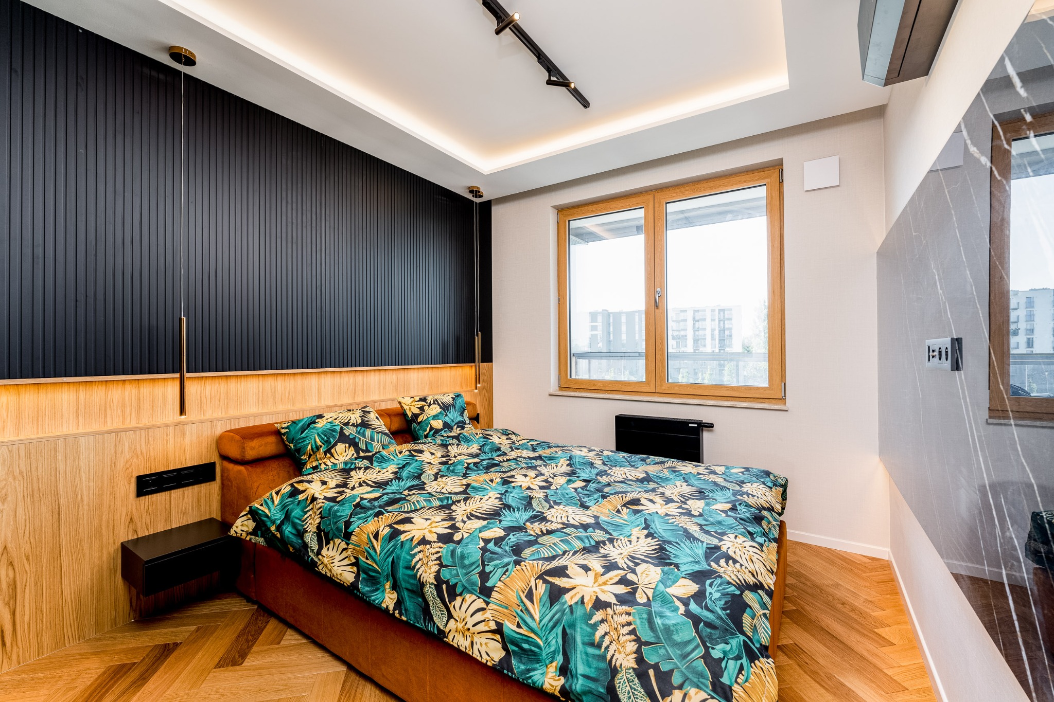 Nowoczesna sypialnia z łóżkiem w tropikalne wzory, drewnianą podłogą w jodełkę i czarną ścianą z pionowymi panelami. Okno z widokiem na miasto.