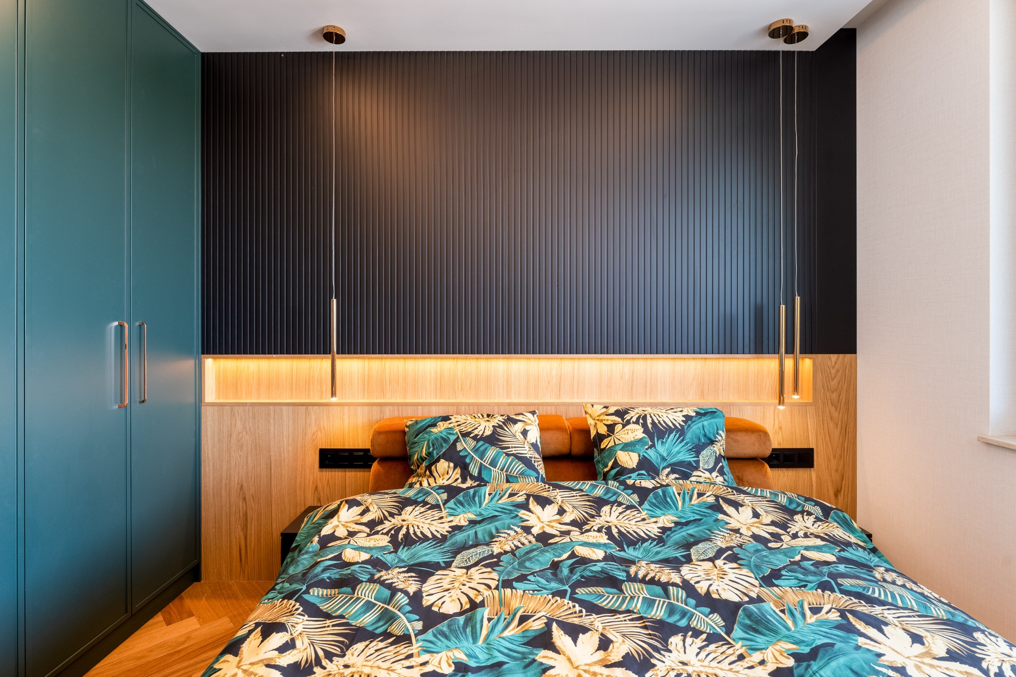 Nowoczesna sypialnia z turkusową szafą, panelem ściennym w kolorze antracytu i drewnianym wezgłowiem z oświetleniem LED. Pościel w tropikalne liście.