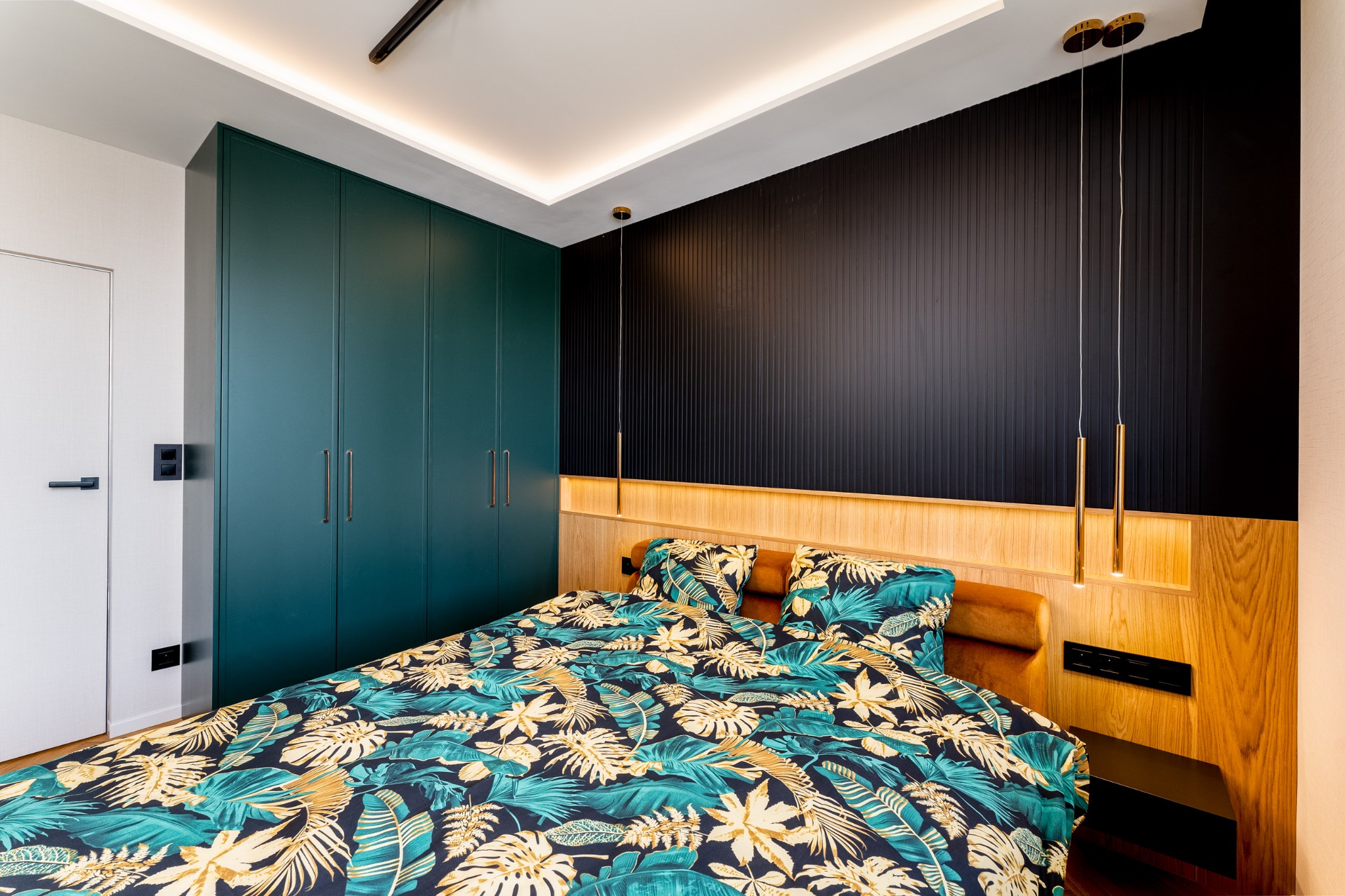 Nowoczesna sypialnia z szafą w zabudowie w odcieniach zieleni, drewnianym wezgłowiem i czarną ścianą z pionowymi panelami. Nad łóżkiem wiszące, złote lampy.