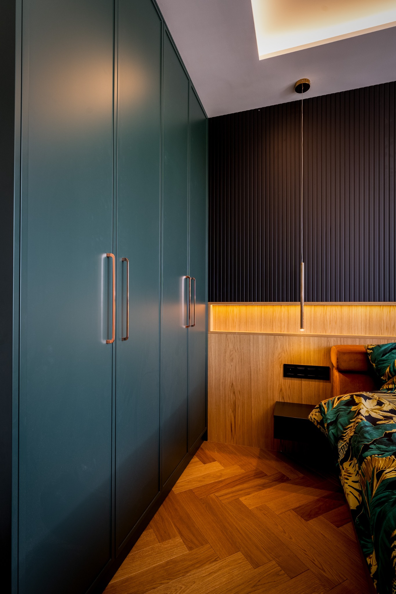 Gładka, ciemnozielona szafa wnękowa z miedzianymi uchwytami w sypialni z drewnianą podłogą w jodełkę i czarną ścianą z lameli.
