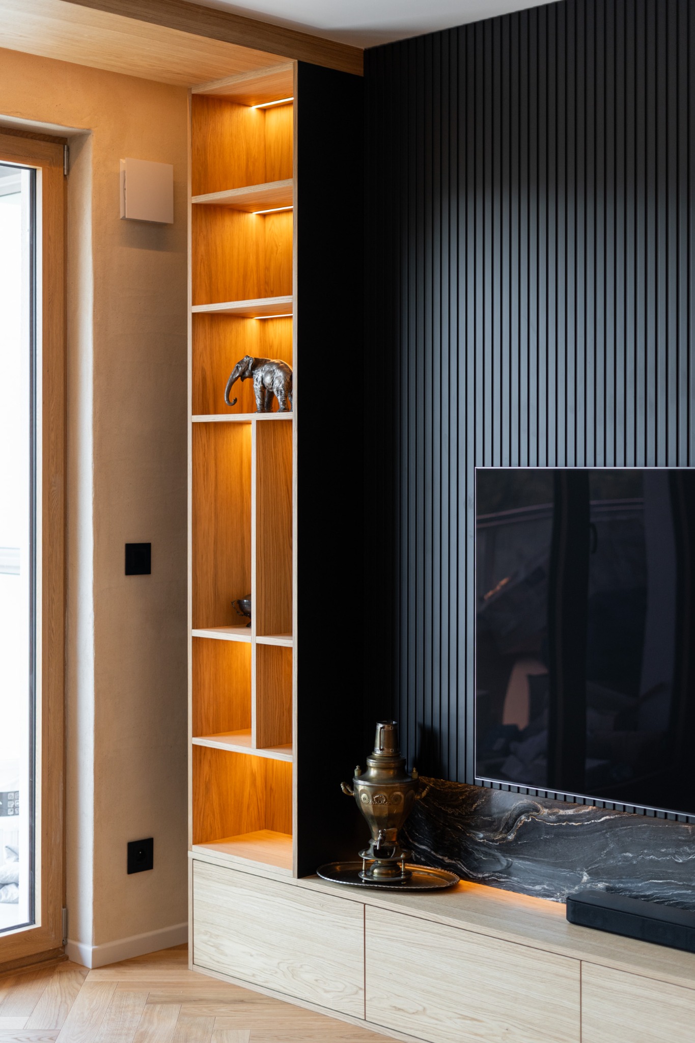 Nowoczesna szafka RTV z podświetleniem LED i otwartym regałem, dekorowana figurką słonia i samowarem. Czarny panel ścienny z pionowymi listwami. Jasne drewno i marmur.