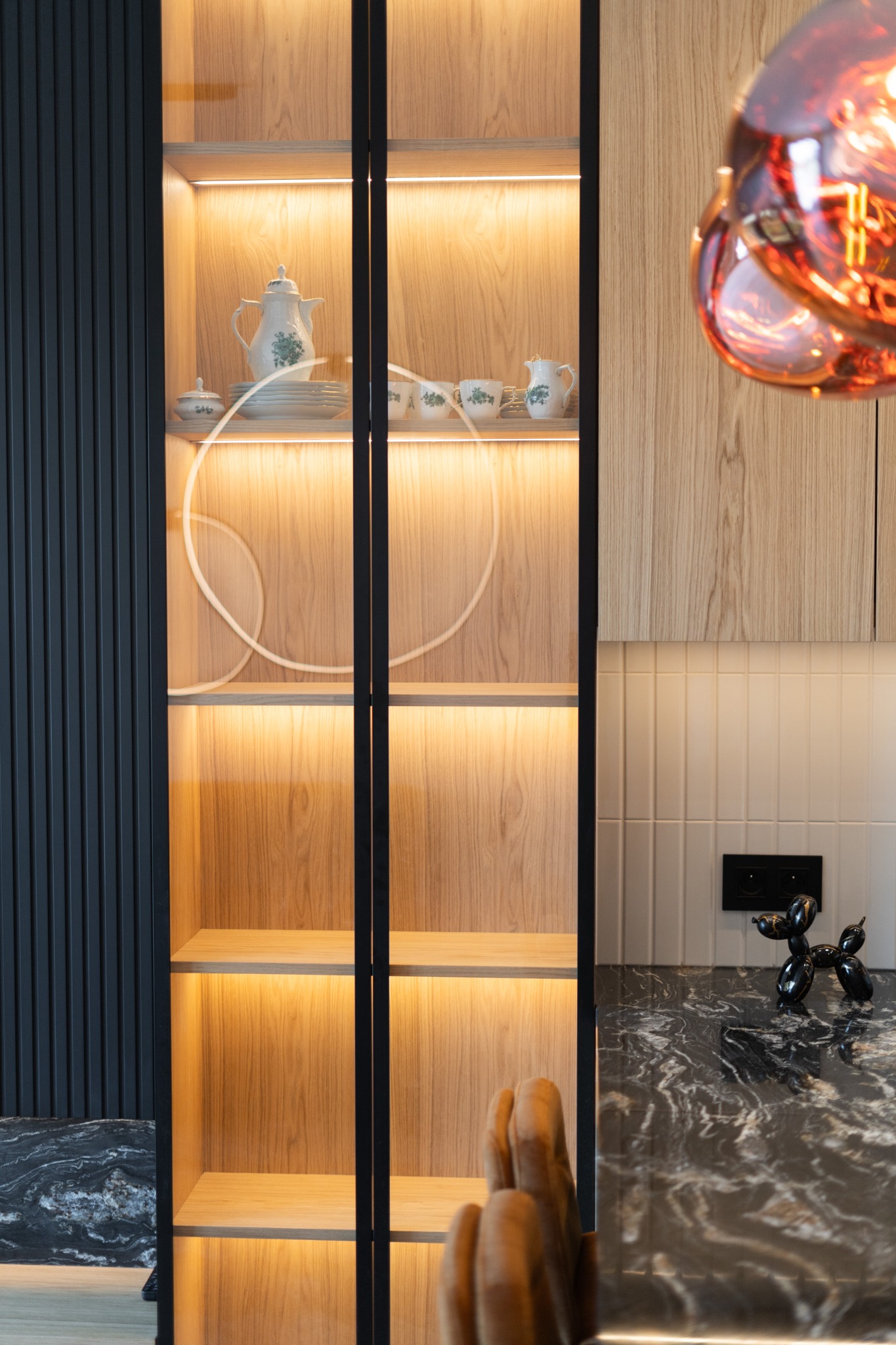 Oświetlona witryna z jasnego drewna z czarnymi ramami, część zabudowy kuchennej. Na półkach porcelana i dekoracyjne oświetlenie. Marmurowy blat i designerska figurka.