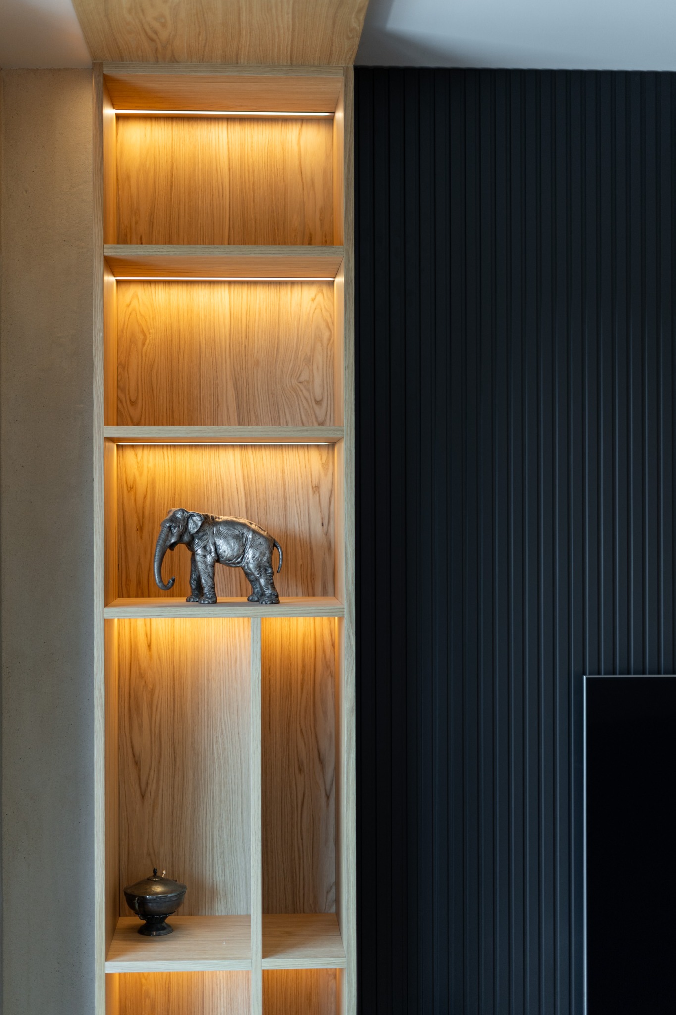 Drewniana szafka wnękowa z oświetleniem LED, dekorowana figurką słonia i ozdobną urną. Ciemna, pionowo żłobiona ściana w tle. Nowoczesny design.
