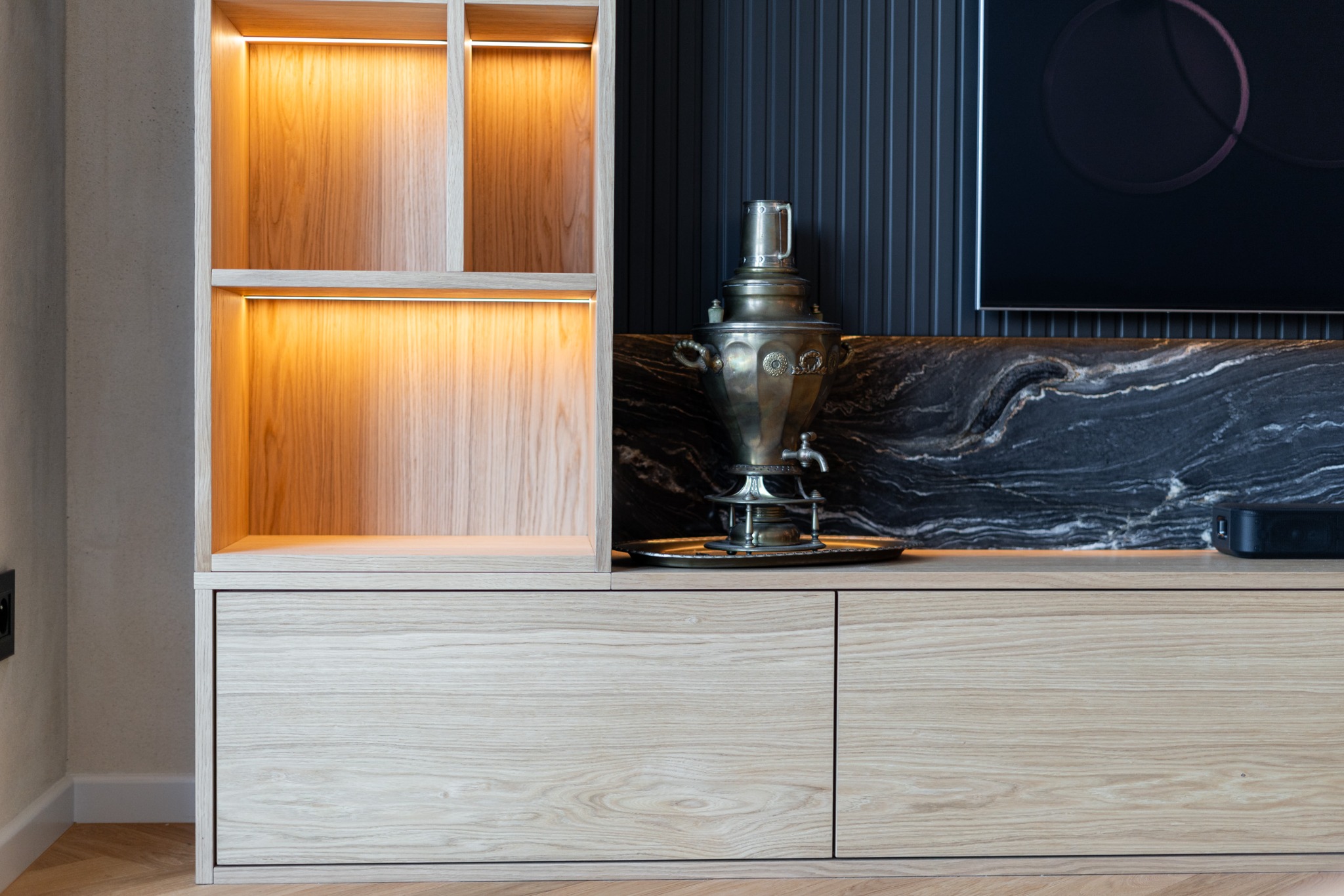 Jasne drewniane meble na wymiar z oświetleniem LED, na blacie samowar na tle czarnego marmuru i telewizora. Minimalistyczny design.
