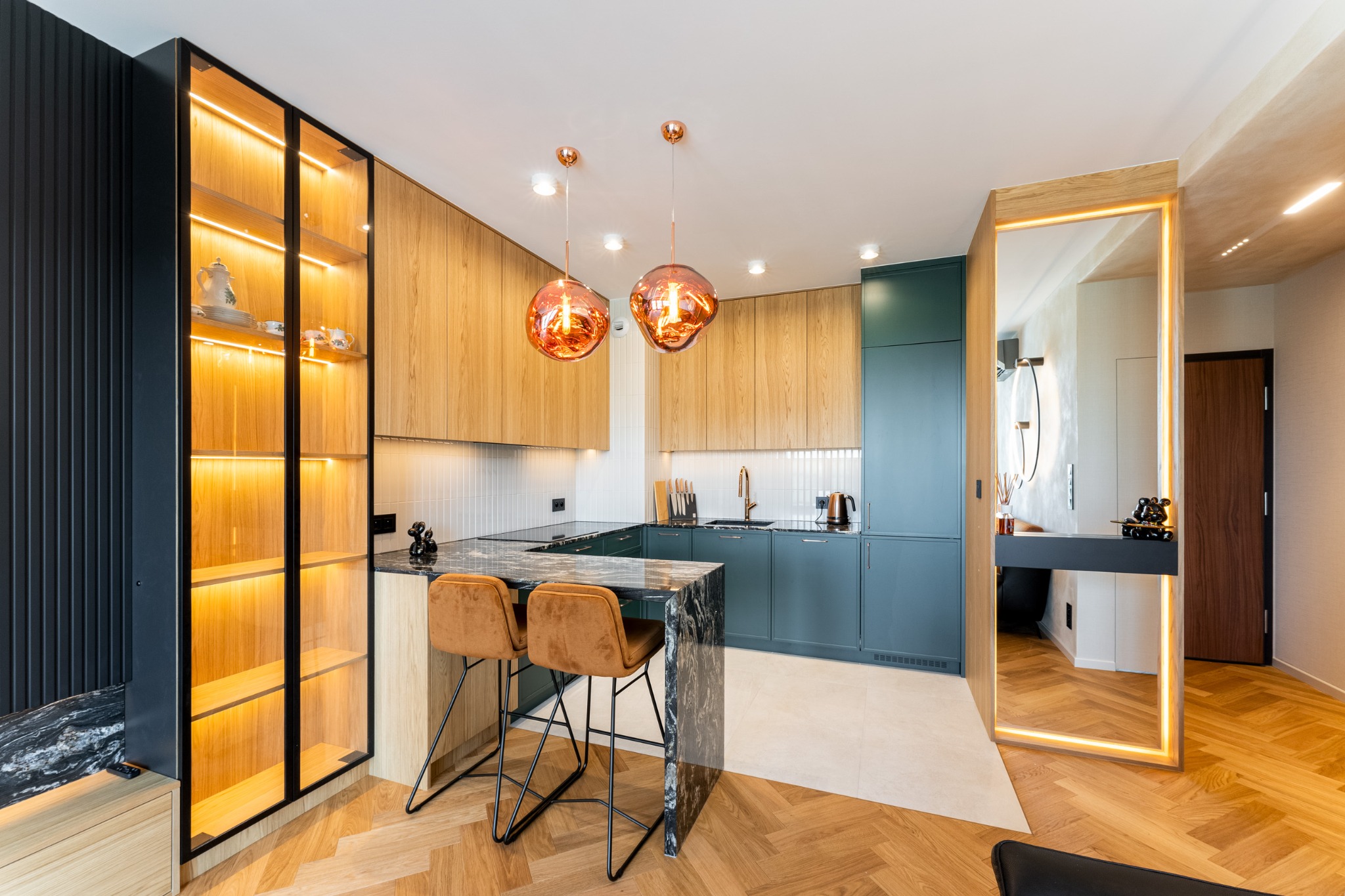 Nowoczesna kuchnia z drewnianymi frontami, zielonymi szafkami i wyspą z marmurowym blatem. Oświetlenie LED podkreśla detale. Dwie miedziane lampy wiszące nad wyspą.